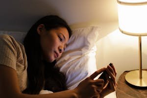 A woman lying in bed at night using a smartphone, illuminated by a bedside lamp.