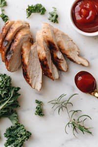 Delicious grilled chicken breast slices served with fresh herbs and ketchup on a marble surface.