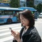 A woman checking her smartphone at a crosswalk in Seoul, where many apps ask for Korean phone number verification
